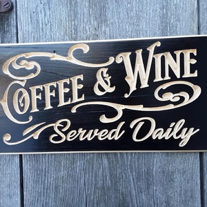May include: A rectangular wooden sign with the words "COFFEE & WINE Served Daily" carved in a light color against a black background. The sign has decorative flourishes and is mounted on a weathered wooden surface.