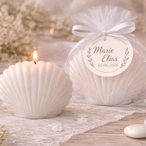 Puede incluir: Dos velas blancas con forma de concha. Una vela está encendida y la otra está envuelta en una bolsa de tul blanco con una cinta blanca y una etiqueta redonda con el texto Marie Elias y la fecha 02.06.2026, rodeadas de detalles florales blancos.
