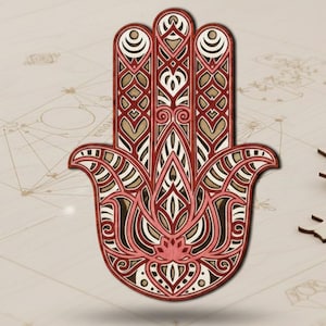 Könnte beinhalten: Ein mehrschichtiges Hamsa-Hand-Kunstwerk aus Holz mit komplizierten geometrischen Mustern in Rot-, Weiß- und Beigetönen. Das Design zeigt ein zentrales Blumenmotiv und ist vor einem hellen Hintergrund platziert. Einige unmontierte Holzstücke sind ebenfalls sichtbar.