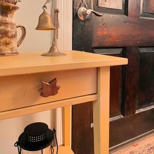 May include: A small, yellow side table with a drawer featuring a leaf-shaped handle. A brass bell sits atop the table, next to a ceramic vase holding dried lavender. A black lantern is below the table, and a dark wooden door is in the background.