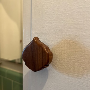 May include: A wooden cabinet knob with a unique, organic shape, possibly resembling a garlic clove. The knob is made of a dark, rich wood, and is mounted on a white cabinet door. The design adds a touch of natural elegance to the cabinetry.
