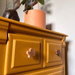 May include: Mustard yellow dresser with two drawers, featuring wooden flower-shaped knobs. A potted plant with a peach-colored pot sits on top. The dresser has a classic design with framed drawer fronts.