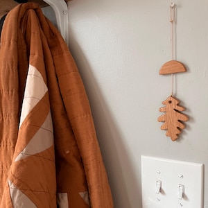 May include: A wooden leaf and crescent moon mobile hangs from a white wall. The mobile is made of natural wood and has a rustic, handmade look. A brown quilted blanket hangs on a hook to the left of the mobile.