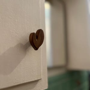 May include: A wooden heart-shaped cabinet knob is attached to a white cabinet door. The knob is a dark brown color and has a three-dimensional design. The background is blurred, showing a glimpse of a green tiled surface.