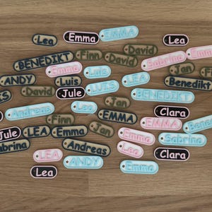 Könnte beinhalten: Eine Sammlung bunter personalisierter Namensschilder mit verschiedenen Namen, darunter Emma, Lea, Luis, Andy, Clara, Sabrina, Jan, Finn, David, Jule und Benedikt.