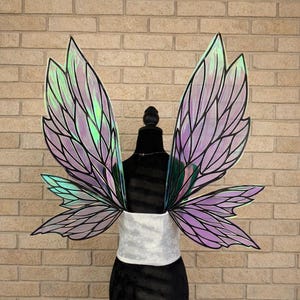 May include: Iridescent fairy wings with a black outline and a purple and green color scheme. The wings are attached to a black mannequin with a white sash. The background is a brick wall.