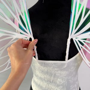 May include: White lace corset top with matching iridescent fairy wings. The wings have a white frame with a rainbow sheen. The corset has a floral pattern and thin straps. The wings are attached to the top.
