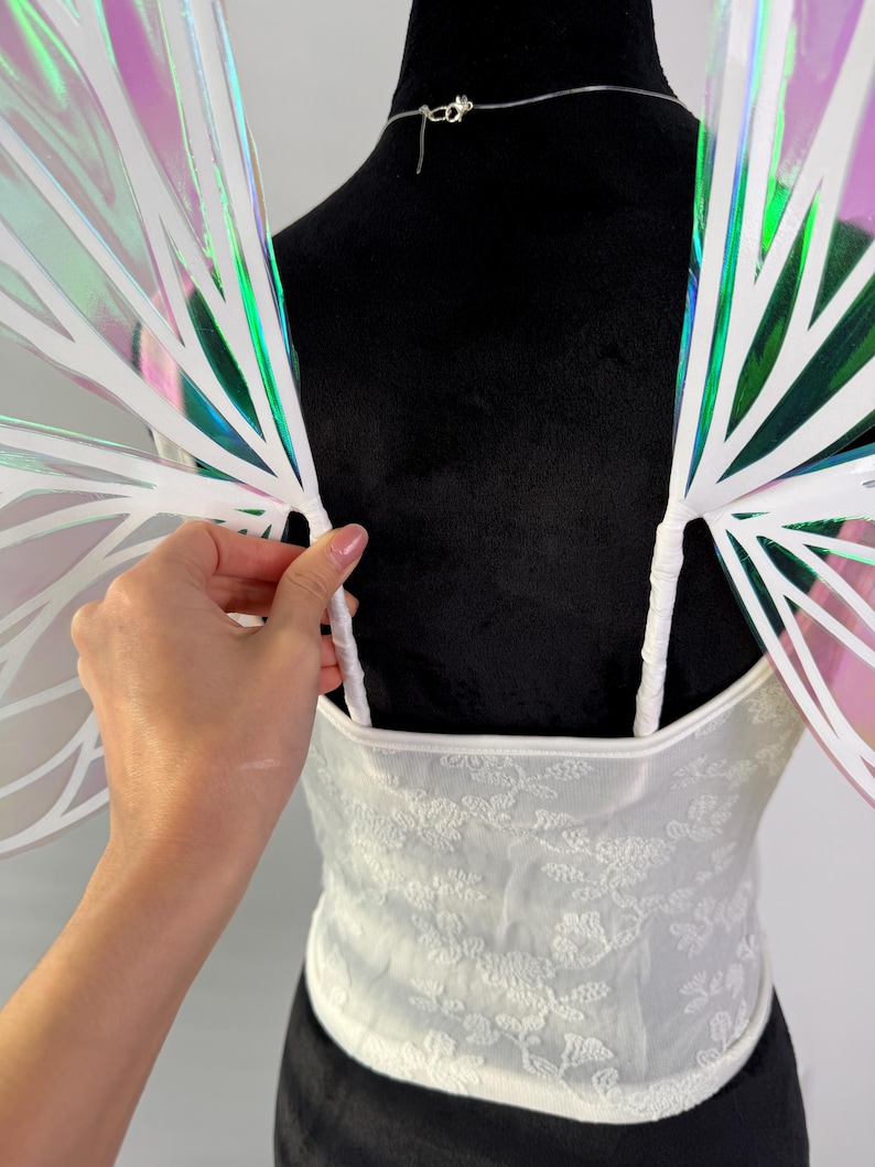 May include: White lace corset top with matching iridescent fairy wings. The wings have a white frame with a rainbow sheen. The corset has a floral pattern and thin straps. The wings are attached to the top.