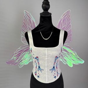 May include: White corset with floral embroidery in blue and red, displayed on a black mannequin. Iridescent fairy wings in shades of purple and green are attached to the mannequin. A pearl necklace is also displayed.