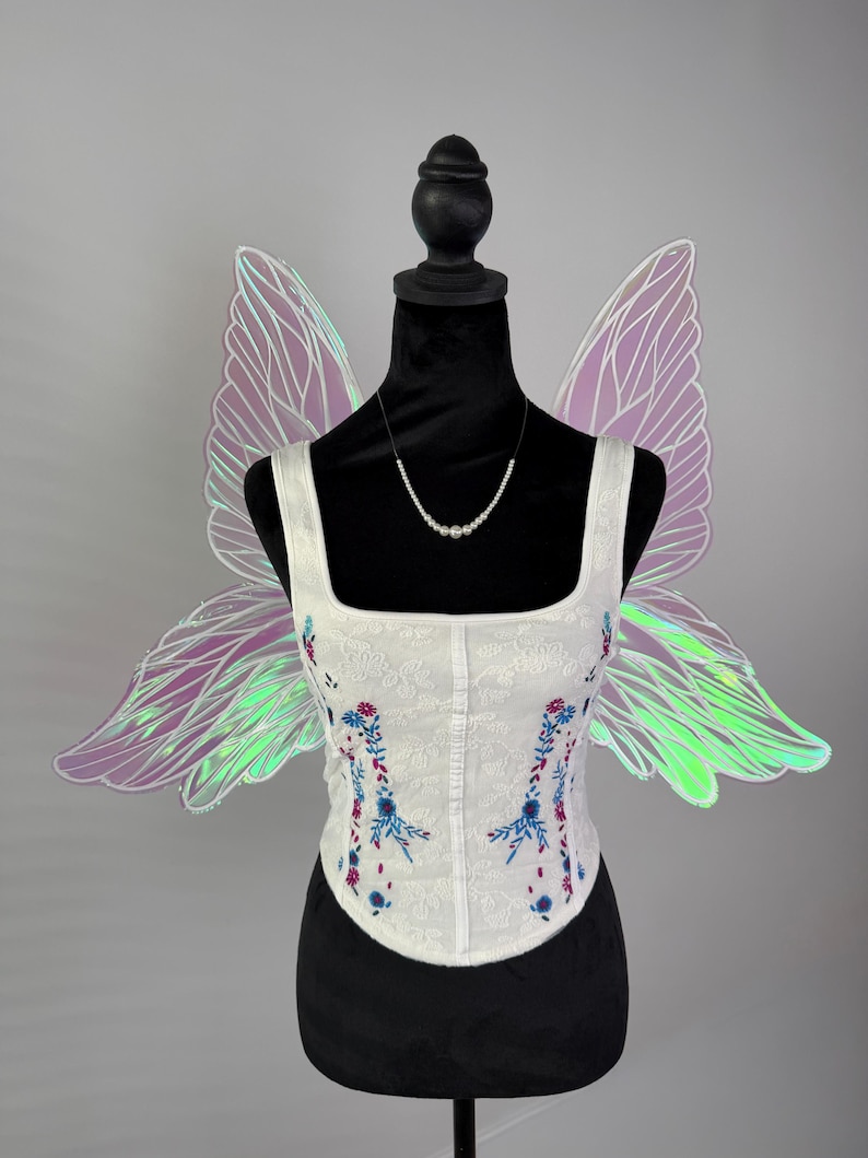 May include: White corset with floral embroidery in blue and red, displayed on a black mannequin. Iridescent fairy wings in shades of purple and green are attached to the mannequin. A pearl necklace is also displayed.
