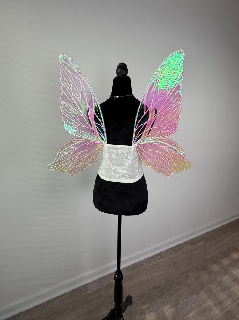 May include: Iridescent fairy wings with a white lace bodice, displayed on a black mannequin. The wings have a delicate, veined design with a gradient of pink, green, and yellow hues. The mannequin is on a black stand.