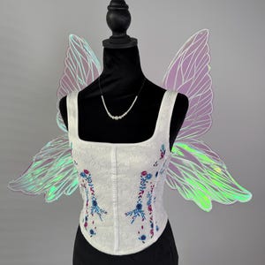 May include: A white corset with floral embroidery in blue and red, displayed on a black mannequin. Iridescent fairy wings in shades of purple and green are attached to the mannequin. A delicate pearl necklace completes the ensemble.