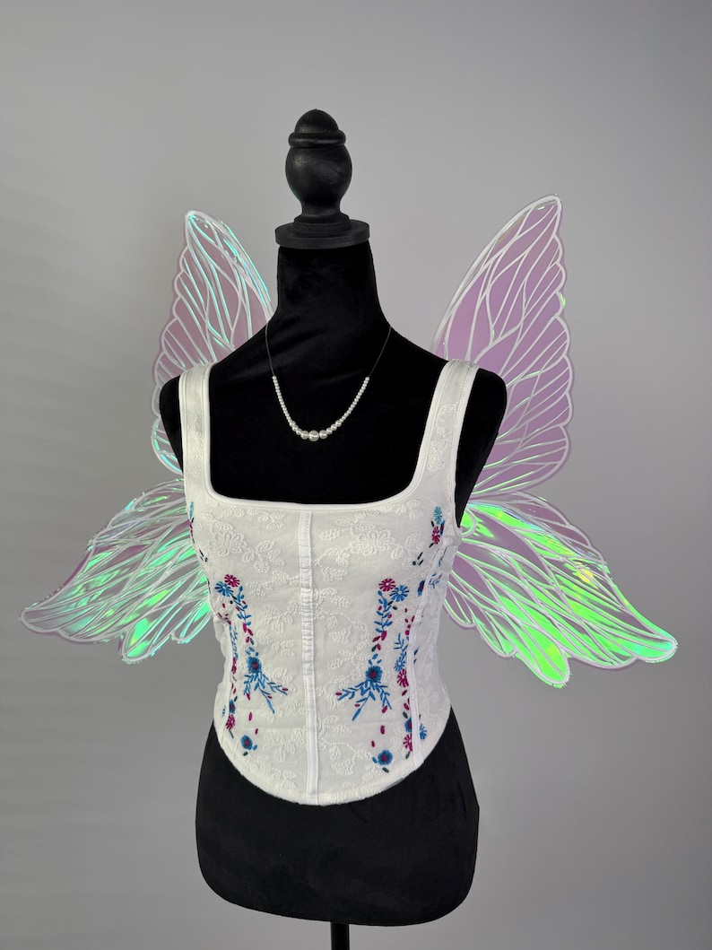 May include: A white corset with floral embroidery in blue and red, displayed on a black mannequin. Iridescent fairy wings in shades of purple and green are attached to the mannequin. A delicate pearl necklace completes the ensemble.