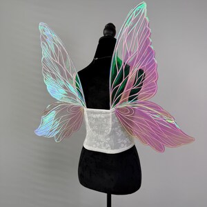 May include: Iridescent fairy wings with a white lace corset. The wings have a gradient of colors, including pink, green, and blue. The wings are attached to a black mannequin. The corset is white with a floral pattern.