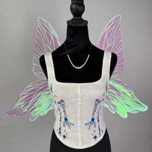 May include: A white corset top with floral embroidery in blue and pink, displayed on a black mannequin. Iridescent fairy wings in shades of purple and green are attached to the mannequin. A pearl necklace completes the ensemble.