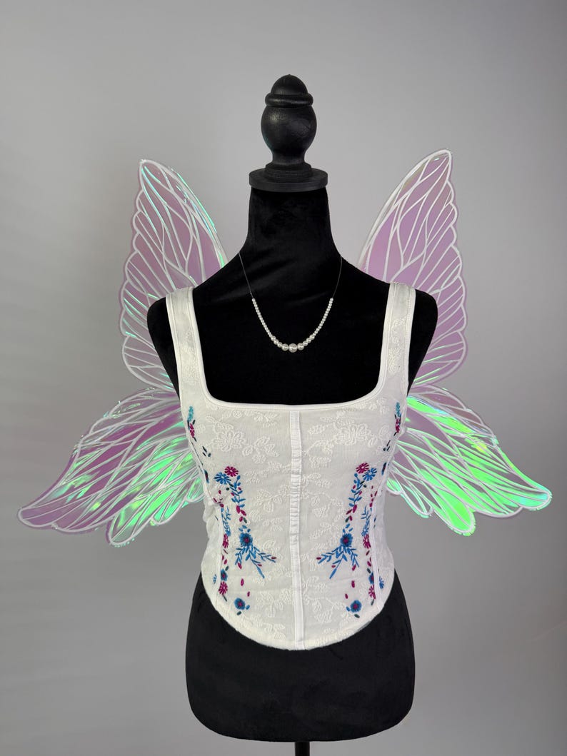 May include: A white corset top with floral embroidery in blue and pink, displayed on a black mannequin. Iridescent fairy wings in shades of purple and green are attached to the mannequin. A pearl necklace completes the ensemble.