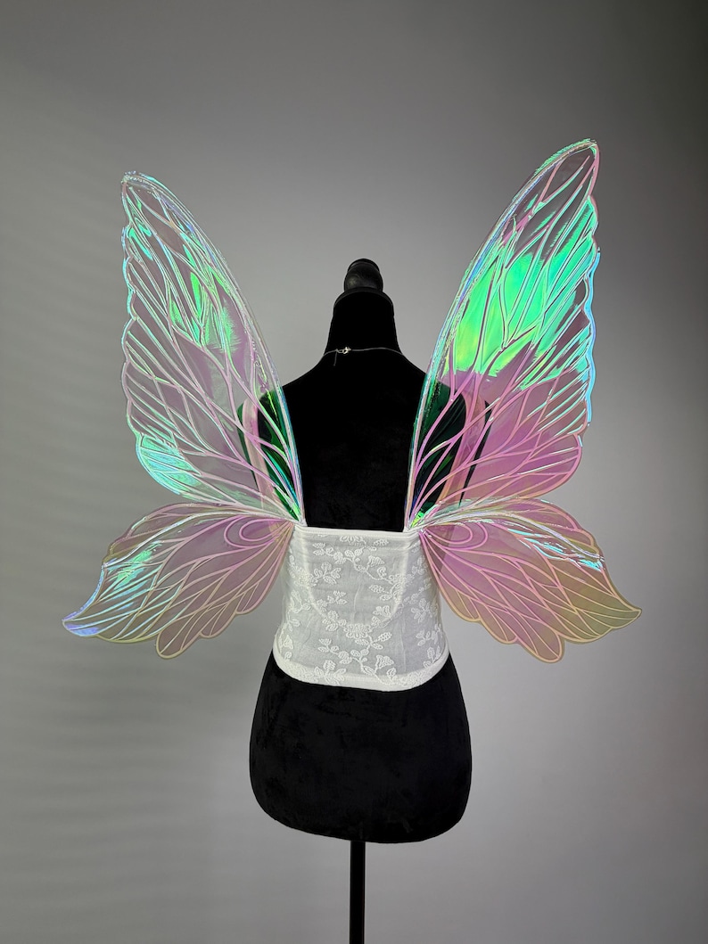 May include: Iridescent fairy wings with a white lace bodice. The wings display a gradient of colors, including pink, green, and yellow. The wings are attached to a black mannequin, showcasing the design and structure of the costume accessory.
