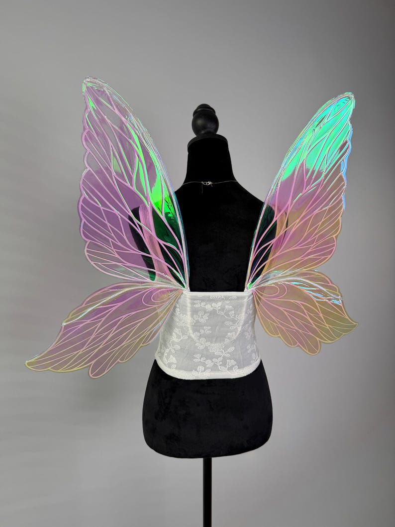 May include: Iridescent fairy wings with a white floral corset. The wings feature a gradient of pink, green, and yellow hues. The corset is made of white lace. The wings are attached to a black mannequin.