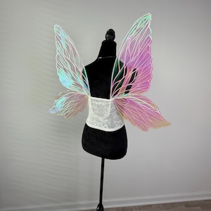 May include: Iridescent fairy wings with a white lace corset are displayed on a black mannequin. The wings have a gradient of colors, including pink, green, and blue. The corset is white with a floral pattern. The mannequin is on a black stand.