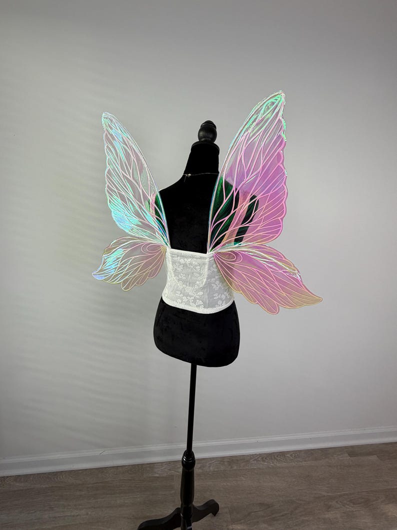 May include: Iridescent fairy wings with a white lace corset are displayed on a black mannequin. The wings have a gradient of colors, including pink, green, and blue. The corset is white with a floral pattern. The mannequin is on a black stand.