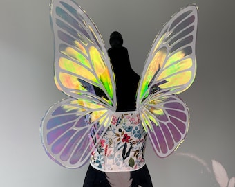 May include: Iridescent butterfly wings with a floral bodice. The wings have a clear, shimmering appearance with a rainbow effect. The bodice is white with a colorful floral pattern. The wings are attached to a black mannequin.