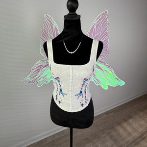 May include: A white corset with floral embroidery and a square neckline is displayed on a black mannequin. Iridescent fairy wings are attached to the mannequin's back. A delicate pearl necklace is also displayed.