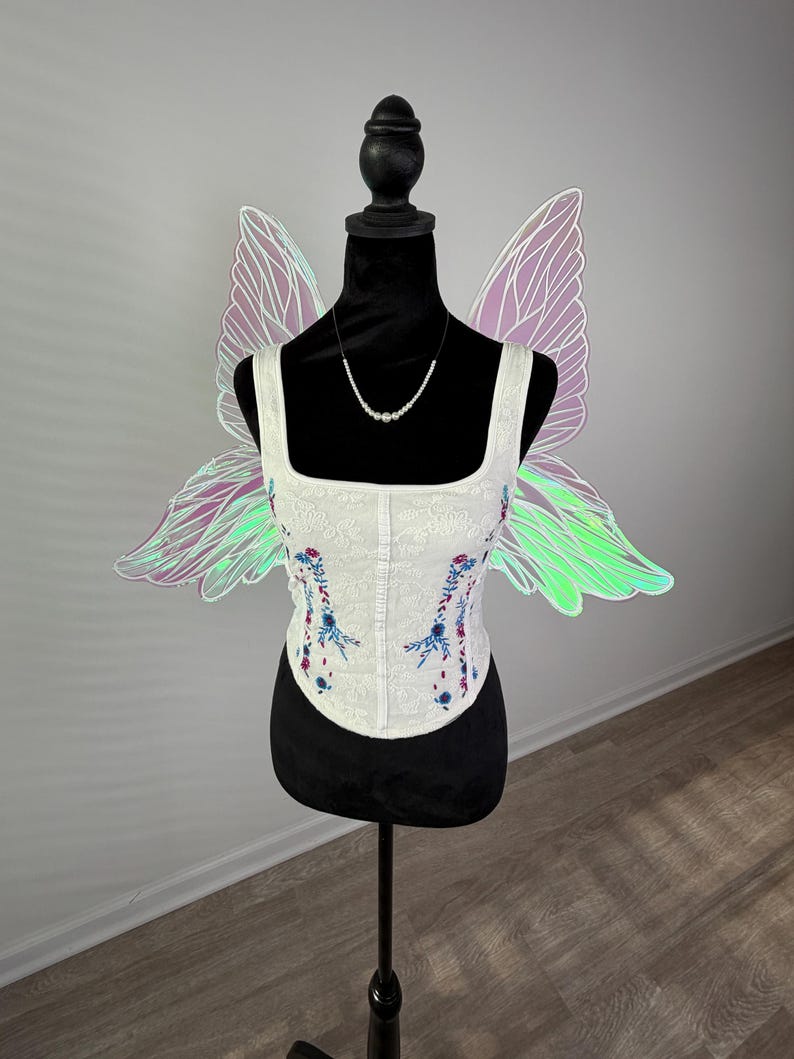 May include: A white corset with floral embroidery and a square neckline is displayed on a black mannequin. Iridescent fairy wings are attached to the mannequin's back. A delicate pearl necklace is also displayed.