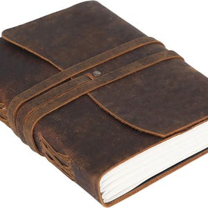 Könnte beinhalten: Ein dunkelbraunes Lederjournal mit einem umlaufenden Riemenverschluss, der durch einen Metallniet gesichert ist. Der Einband hat eine abgenutzte Textur und der Buchrücken zeigt eine dekorative Lederschnürung. Das Notizbuch enthält weiße Papierseiten.