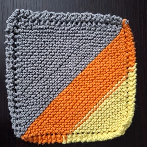 May include: A square, hand-knitted washcloth with a diagonal color block design. The top left is gray, the center is orange, and the bottom right is yellow. The edges are finished with a textured border.