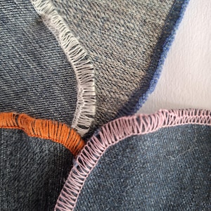 Può includere: Diversi strati di tessuto denim blu con bordi tagliacuci a vista. Le cuciture sono rifinite con punti overlock in fili di colori contrastanti, tra cui bianco, arancione brillante e rosa chiaro, che mettono in risalto la costruzione tessile.