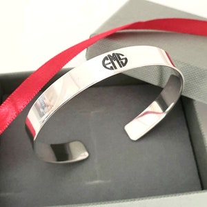 May include: A silver cuff bracelet with the initials "EMS" engraved on the top. The bracelet is presented in a gray gift box with a red ribbon. The bracelet has a polished, reflective surface.