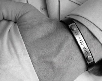 May include: Black leather bracelet with a silver plate engraved with Roman numerals. The bracelet is secured with two silver rivets. The image is in black and white, showing the bracelet on a person's wrist, with a light-colored shirt sleeve in the background.