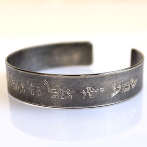 May include: A silver cuff bracelet with engraved Hebrew script. The bracelet has a brushed metal finish and a simple, open design. The text appears to be a religious inscription.
