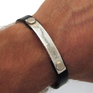 May include: A black leather bracelet with a silver band engraved with a pattern. The band has two silver snap closures.