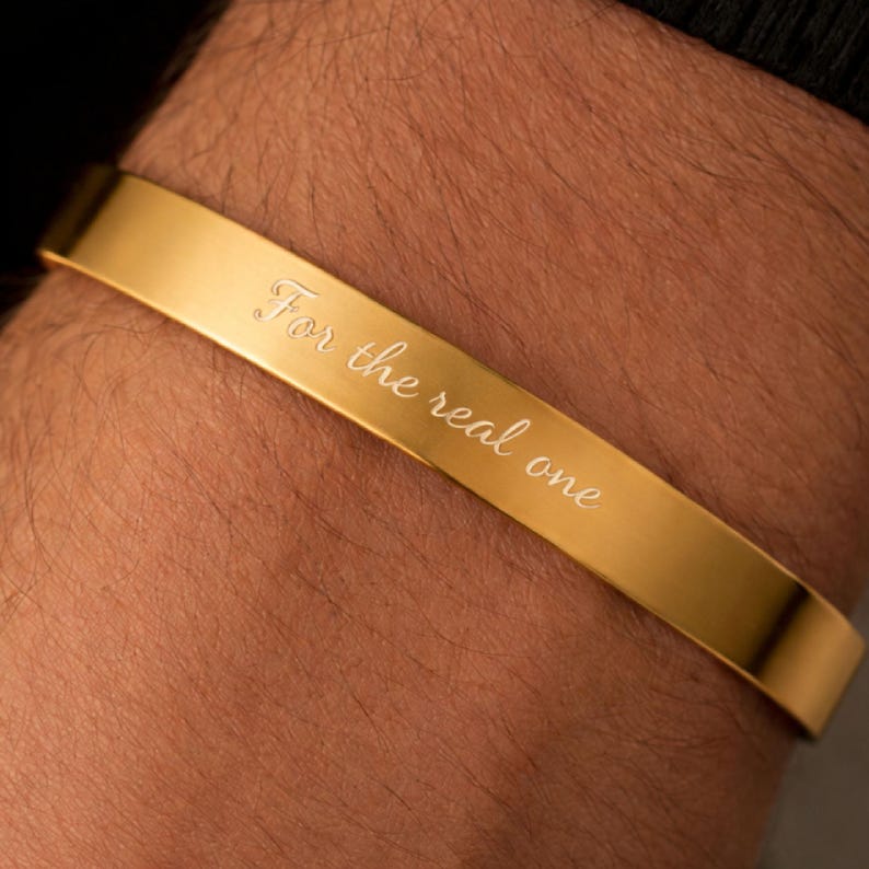 May include: A gold-toned cuff bracelet with a smooth, polished finish. The surface of the bracelet features the engraved text "For the real one" in a cursive script font. The piece is a simple, open-ended band design with a consistent width.
