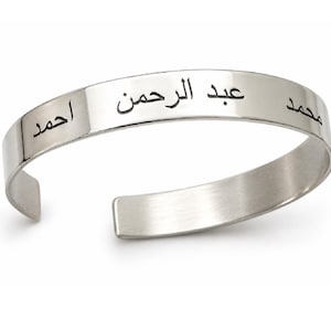 May include: A silver-toned metal cuff bracelet with an open back design. The outer surface features engraved black Arabic script text that reads "عبد الرحمن" in the centre, flanked by the names "احمد" on the left and "محمد" on the right side.