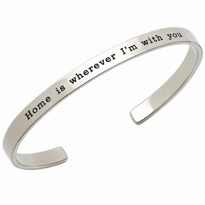May include: A silver-colored cuff bracelet with the engraved text "Home is wherever I'm with you." The bracelet has a simple, elegant design and is curved to fit the wrist.