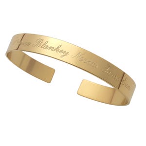 May include: A gold-toned metal cuff bracelet with an open back design. The outer surface features an engraved inscription in a cursive script that reads Prince Blankey Nerons Jame Sun. The bracelet has a smooth, polished finish throughout.