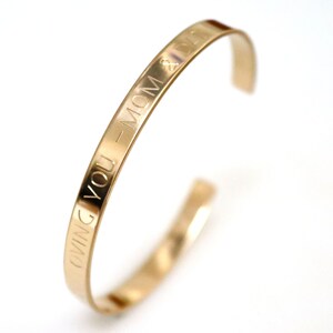 May include: A gold-colored cuff bracelet with the engraved words "LOVING YOU - MOM & DAD". The bracelet has a simple, open design and is set against a white background. The text is clearly visible.