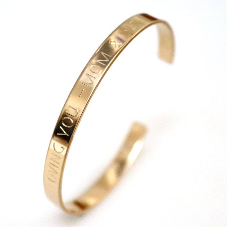 May include: A gold-colored cuff bracelet with the engraved words "LOVING YOU - MOM & DAD". The bracelet has a simple, open design and is set against a white background. The text is clearly visible.