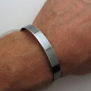 May include: A silver bracelet with a flat, wide band. The bracelet is worn on a person's wrist.