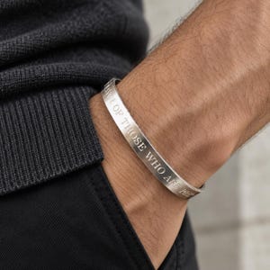 May include: A silver-toned metal cuff bracelet featuring an open design and a smooth, polished finish. The outer surface of the bracelet is engraved with the text "DAWN OF THOSE WHO ARE PRE". The bracelet is worn on a wrist against a dark fabric sleeve.