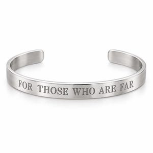 May include: A silver-toned metal cuff bracelet with an open back design. The outer surface of the bracelet features the engraved text FOR THOSE WHO ARE FAR in a clean, capitalised serif font. The metal has a smooth, polished finish throughout.