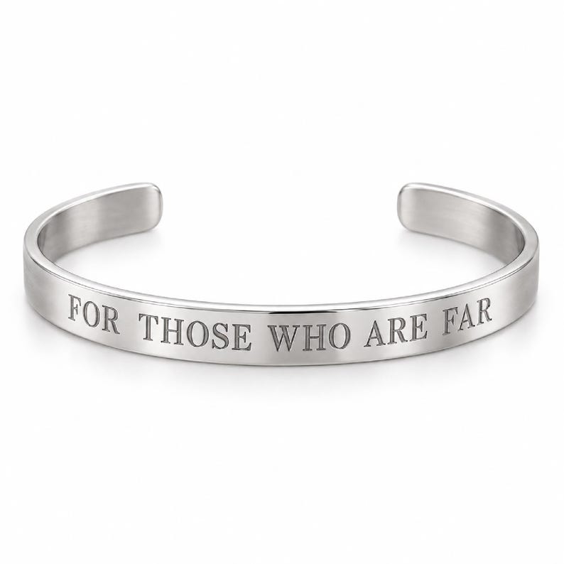 May include: A silver-toned metal cuff bracelet with an open back design. The outer surface of the bracelet features the engraved text FOR THOSE WHO ARE FAR in a clean, capitalised serif font. The metal has a smooth, polished finish throughout.