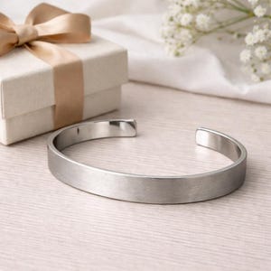 May include: A silver-toned metal cuff bracelet with a brushed finish and an open back design, positioned next to a square gift box featuring a light beige ribbon tied in a bow, showcasing a clean and simple aesthetic for this jewellery piece.