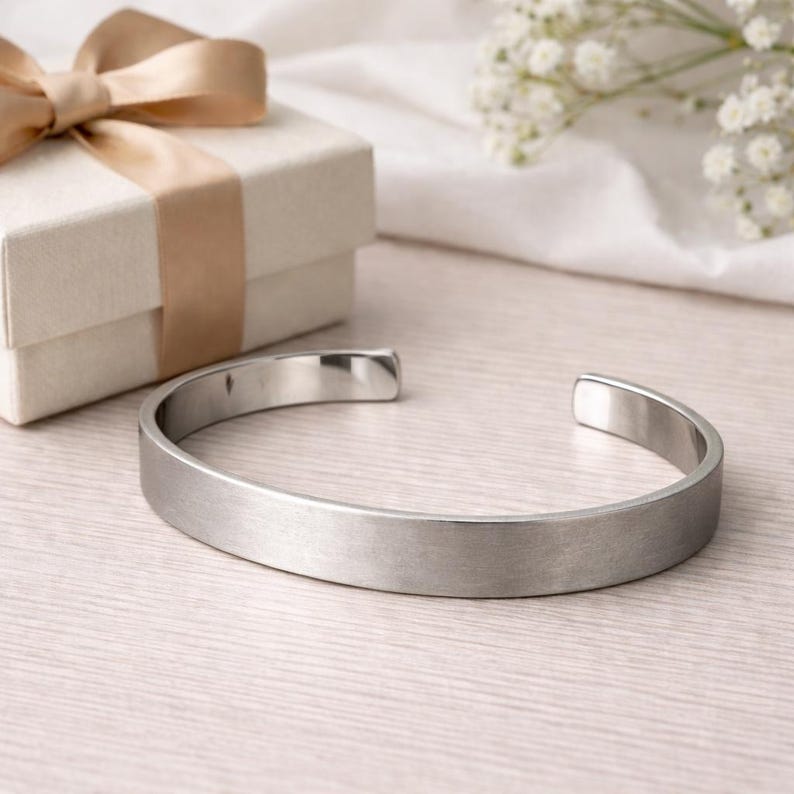 May include: A silver-toned metal cuff bracelet with a brushed finish and an open back design, positioned next to a square gift box featuring a light beige ribbon tied in a bow, showcasing a clean and simple aesthetic for this jewellery piece.