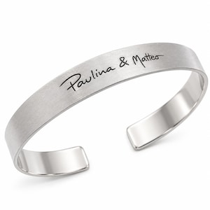 May include: A silver-toned cuff bracelet with a brushed matte finish on the exterior and a polished interior. The outer surface features the black engraved text Paulina & Matteo in a cursive script font, with an open back design for adjustment.