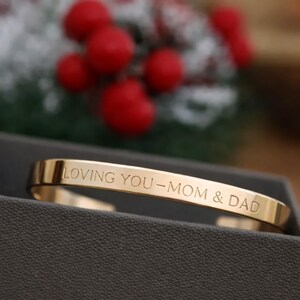 May include: A gold-colored cuff bracelet with the engraved message "LOVING YOU - MOM & DAD." The bracelet is displayed in a dark gray box, with blurred red berries and greenery in the background.