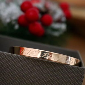 May include: A gold-colored bangle bracelet with engraved text. The bracelet is resting in a gray box, with red berries and greenery in the background. The engraved text on the bracelet reads "KHS Blackbird S".