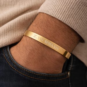 May include: A gold-toned metal cuff bracelet featuring a smooth, polished finish. The outer surface of the bracelet is engraved with the Roman numerals X. VII. MMXXIV in a clean, sans-serif font, centred along the length of the band.